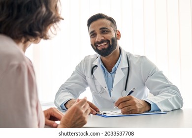 Young cheerful Indian doctor therapist in white robe having appointment with female patient sitting at table filling medical form in modern clinic hospital. Over shoulder view. Healthcare concept.