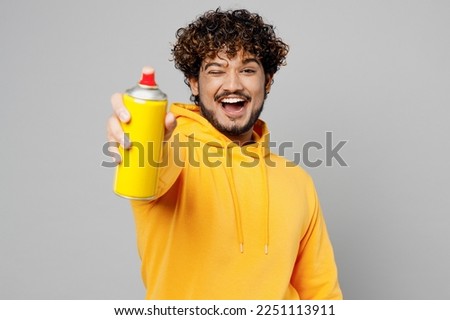 Young cheerful Indian man 20s he wearing casual yellow hoody hold in hand graffiti spray paint bottle point camera on you isolated on plain grey background studio portrait. People lifestyle portrait