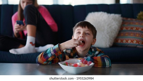 Young cheerful kid eating delicious sugar candies by table in living room checking whether anybody watching. His sister using mobile phone - Shutterstock ID 1955263984
