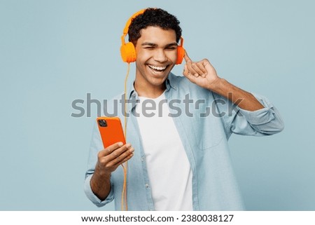 Young cheerful man of African American ethnicity he wears shirt casual clothes listen to music in headphones use mobile cell phone isolated on plain pastel light blue cyan background studio portrait