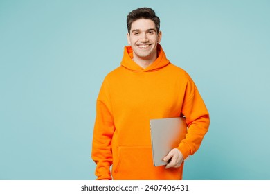 Young cheerful IT man he wears orange hoody casual clothes hold closed laptop pc computer looking camera isolated on plain pastel light blue cyan color background studio portrait. Lifestyle concept - Powered by Shutterstock