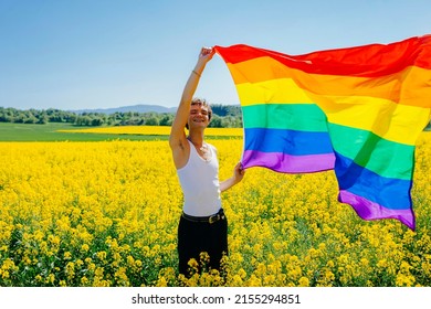 Young Cheerful Man Holding The LGBT Gay Rainbow Flag On Yellow Rapeseed Field In Spring. Happiness, Freedom And Love Concept For Same Sex Couples. Lifestyle Outdoors.
