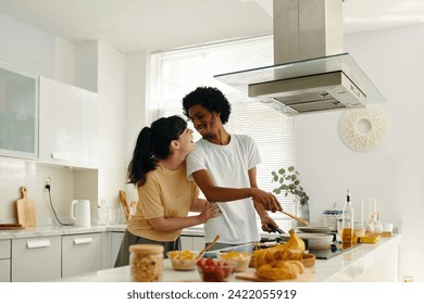 Young cheerful man looking at his wife while standing by electric stove in the kitchen and preparing something tasty for breakfast - Powered by Shutterstock