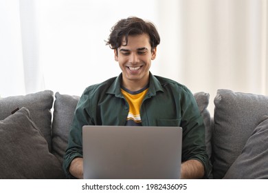 Young cheerful man sitting on sofa with laptop indoors looking at computer monitor and smiling. Handsome young man enjoying watching funny video on social media