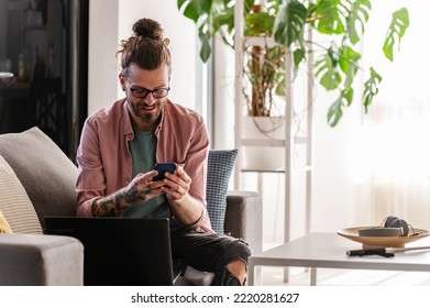 Young Cheerful Man With A Sleeve Tattoo Using Laptop Computer And Smartphone For Paying Bills Online Using Banking App.