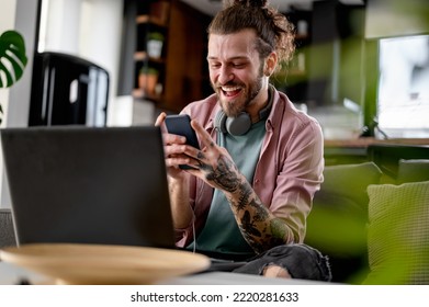 Joven hombre alegre con un tatuaje de manga usando computadora portátil y smartphone. Empresario independiente trabajando desde casa usando aplicaciones bancarias.