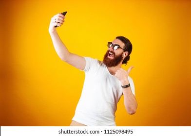 Young Cheerful Man Taking A Selfie On His Smartphone Wearing A White T-shirt And Earpods. Bearded Long Haired Hipster Looking At His Iphone Taking Picture And Showing Thumb Up On Yellow Background. 