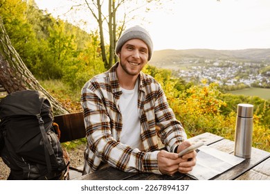 Young cheerful man tourist with backpack using mobile phone while sitting on wooden bench on hillside - Shutterstock ID 2267894137
