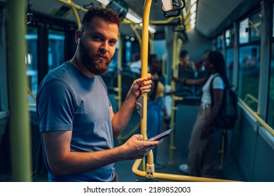 Young cheerful man using a smartphone during his ride during a night and holding onto the bar while standing in a bus. Handsome man taking bus to work. Urban public transportation concept. - Powered by Shutterstock