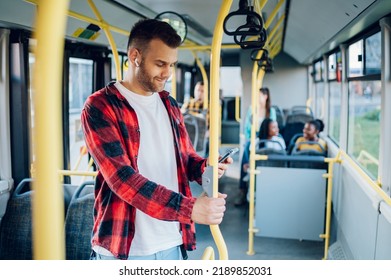 Young cheerful man using a smartphone during his ride and holding onto the bar while standing in a bus. Handsome man taking bus to work while browsing social media. Urban public transportation concept