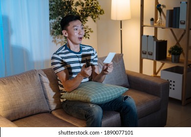 Young Cheerful Man Watching Gambling Game On Television Program In Late Night Hands With Control Remote And Lottery Ticket Sit On Sofa And Looking Thrilled. Shocked Male Surprised By Tv In Evening