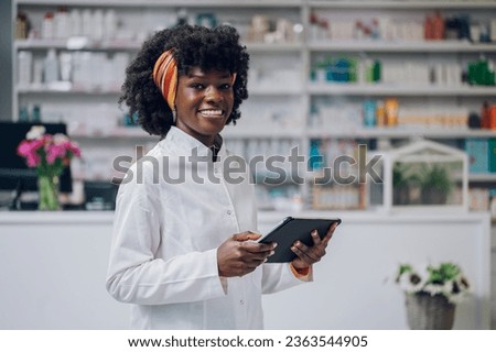 A young cheerful multicultural chemist is standing in a pharmacy with a tablet in her hands and smiling at the camera. A pharma worker is using the tablet to sell healthcare products online.