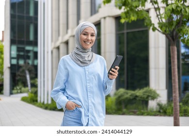 A young, cheerful Muslim woman wearing a hijab, holding a smartphone, walks confidently beside an office building. - Powered by Shutterstock