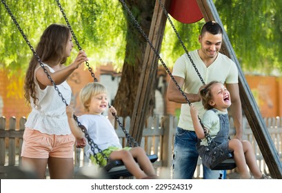 Young cheerful parents with kids at playground's swings. Focus on man 