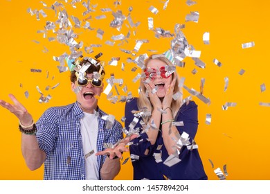 Young cheerful positive couple of funny glasses and a paper cap rejoice and shout on a yellow background with flying confetti. Conception of a holiday and fun. - Shutterstock ID 1457894204