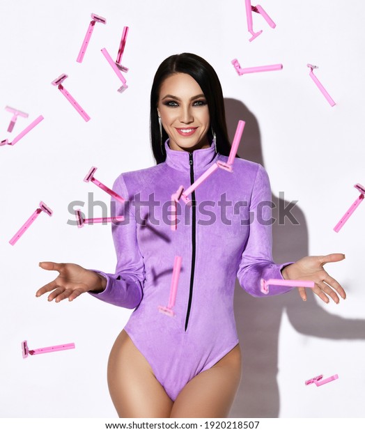 Young cheerful pretty brunette woman in purple\
jumpsuit with long sleeves stands surrounded with falling down\
tossed up pink razors over white background. Laser hair removal,\
epilation concept