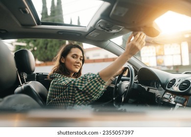 Young cheerful pretty woman, sitting in a car, behind the wheel and adjusting a rear mirror panel and looking at the camera. - Powered by Shutterstock