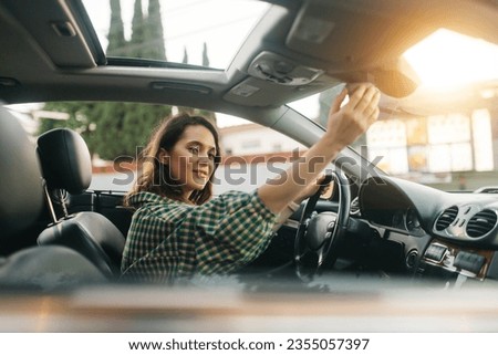 Young cheerful pretty woman, sitting in a car, behind the wheel and adjusting a rear mirror panel and looking at the camera.