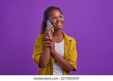 Young cheerful sociable African American woman talking on phone and smiling broadly enjoying calling parents or chatting with friends dressed in oversize shirt stands on plain lilac background - Powered by Shutterstock