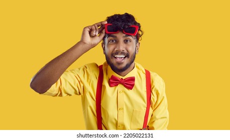 Young Cheerful South Asian Man Raises Funny Sunglasses From Eyes And Looks At Screen With Smile Rejoicing At Unexpected Meeting At Party Or Concert Standing In Yellow Studio. Indian Showman Concept