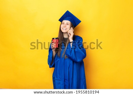 A young cheerful student in graduating uniform is talking to the phone happily and drinking a hot drink .