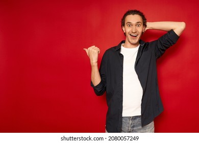 Young Cheerful Surprised Confused Male Student In Casual Blue Shirt Points With His Index Finger To Side Free Work Area Mock Up Of Copy Space Isolated On Red Background Studio Portrait. Promotion