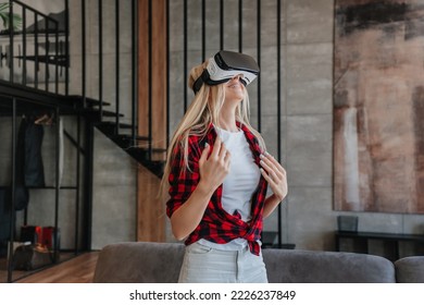 Young Cheerful Swedish Woman In Plaid Shirt And White Pants Using Vr Glasses At Home. Relaxing Swedish Girl In Virtual Reality Headset Standing In Room With Sofa, Stairs, Playing Games. Leisure