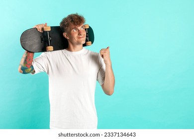 young cheerful tattooed guy in white t-shirt holds skateboard and smiles on blue isolated background, hipster skater shows his hand to the side and advertises copy space - Powered by Shutterstock