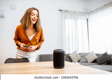 Young cheerful woman controlling home devices with a voice commands, talking to a smart column at home. Concept of smart home and voice command control - Powered by Shutterstock