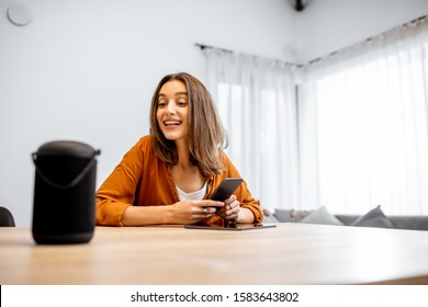 Young Cheerful Woman Controlling Home Devices With A Voice Commands, Talking To A Smart Column At Home. Concept Of Smart Home And Voice Command Control