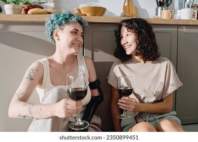 Young cheerful woman with disability and her girlfriend toasting with glasses of red wine while sitting by sunlit kitchen counter in the morning - Powered by Shutterstock