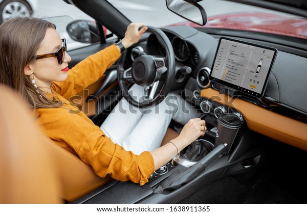 Young and cheerful woman driving sports car\
with a digital touchscreen with launched controlling program on the\
front dashboard