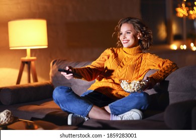 Young  Cheerful Woman Eating Popcorn And Watches  Movie On  Cable TV While Switching Channels With The Remote Control At Home In Evening  Alone
