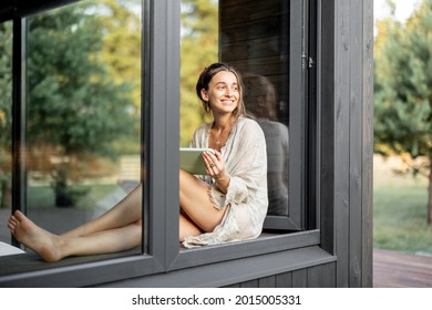 Young Cheerful Woman Enjoy Of Resting At Modern House Or Hotel, Sitting With Tablet On The Window Sill And Looking At Pine Forest. View From Outside. Concept Of Solitude And Recreation On Nature.