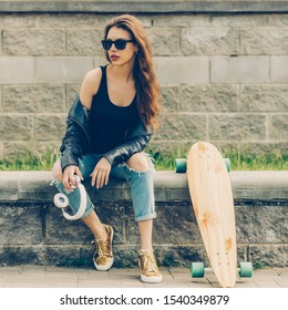 Young cheerful woman in fashion sunglasses and rock black style leather jacket with longboard skateboard and white music headphones. Hipster urban style girl. Lifestyle outdoor city portrait.