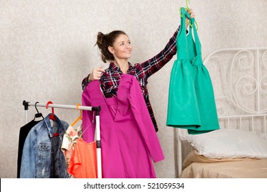 Young cheerful woman holding two colorful bright dresses and choosing what to wear. Happy girl getting ready for special event or date and picking apparel in her bedroom from clothes on the rack