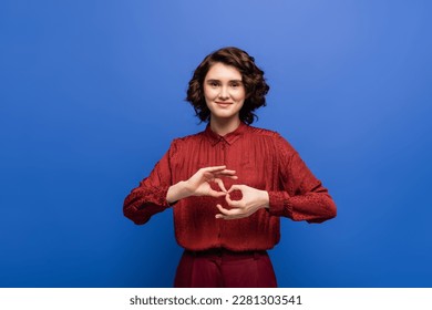 young and cheerful woman looking at camera and showing symbol meaning interpreter on sign language isolated on blue