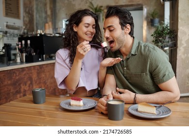 Young cheerful woman looking at her boyfriend eating piece of tasty cheesecake and having cappuccino during romantic date in cafe - Powered by Shutterstock