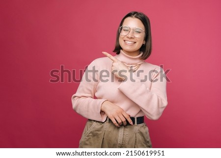 Young cheerful woman is pointing aside with a finger to the copy space. Studio shot over pink background.