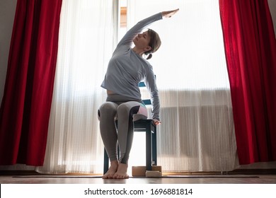 Young Cheerful Woman Practicing Yoga, Sitting On Chair Doing Side Bend Exercise, Working Out At The Living Room At Home