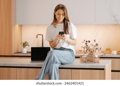 Young cheerful woman is sitting on countertop while scrolling her phone in the kitchen. - Powered by Shutterstock