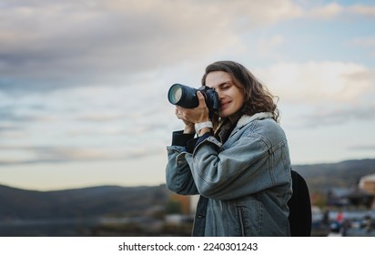 Young cheerful woman traveler in a denim jacket with a backpack holds a camera in her hands while standing on the shore of the mountain  lake - Powered by Shutterstock