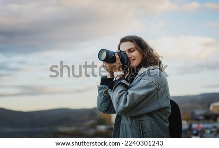 Young cheerful woman traveler in a denim jacket with a backpack holds a camera in her hands while standing on the shore of the mountain  lake