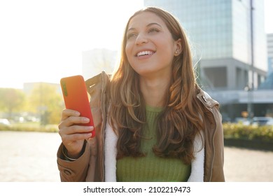 Young cheerful woman using smartphone over city background smiling looking to the side and staring away thinking