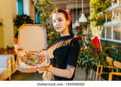 Young Cheerful Woman Waiter Wearing Black T-shirt Holding Freshly Baked Pizza In Open Box At Pizza Shop Looking At Camera With Modern Interior Of Pizza Shop Or Cafeteria.