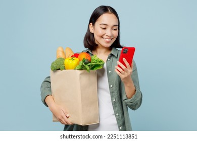 Young cheerful woman wear casual clothes hold brown paper bag with food products use mobile cell phone isolated on plain blue cyan background studio portrait. Delivery service from shop or restaurant - Powered by Shutterstock