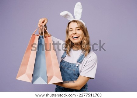 Young cheerful woman wears casual clothes bunny rabbit ears hold paper package bags after shopping isolated on plain light pastel purple background studio portrait. Happy Easter sale buy day concept