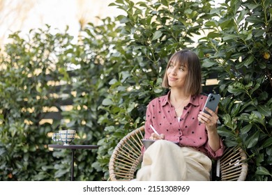 Young cheerful woman works on smart phone and tablet while sitting on background of green bushes outdoors. Concept of remote work at cozy garden
