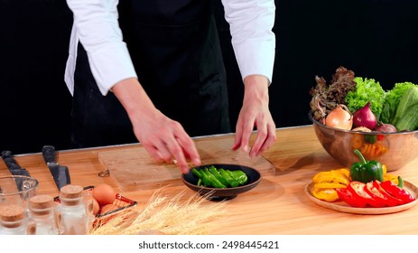 Young chef in black apron carefully plates sliced green bell pepper in bowl, surrounded by fresh vegetables and ingredients - Powered by Shutterstock