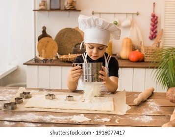 young chef in a white apron and cap sifts flour into dough at a wooden table in the kitchen